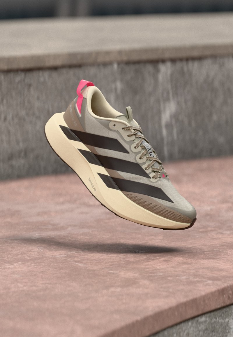Ανοιχτό μπεζ αθλητικό παπούτσι με μαύρες ρίγες, μπεζ σόλα και ροζ ταμπελάκι στη φτέρνα που αιωρείται πάνω από μια ροζ τσιμεντένια επιφάνεια.