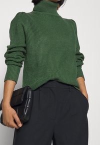 Pull vert en maille avec un col roulé haut, une texture côtelée et des manches bouffantes, associé à un pantalon noir sur mesure et une pochette noire matelassée.