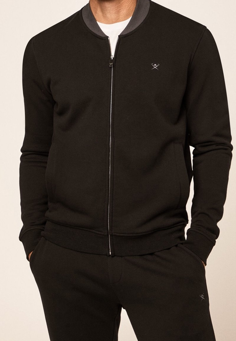 Svart zip-up sweatshirt med grå krage, ribbad mudd och nederkant. Har en liten logotyp på bröstet och en mjuk tygkvalitet.