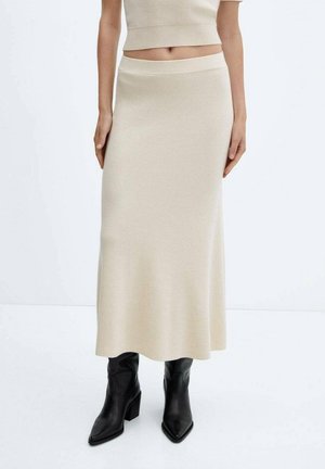 Femme portant une jupe midi côtelée beige à taille haute et des bottes à talons pointues noires sur fond blanc.