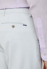 Hellgraue Hose mit einer Gesäßtasche, ausgestattet mit einem metallischen Knopf und einem kleinen Markenetikett. Ein hellviolettes Hemd ist teilweise zu sehen.