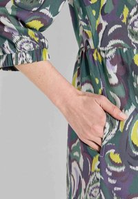 Hand ruht in der Seitentasche eines Kleidungsstücks mit einem bunten abstrakten Muster in Grün, Lila, Gelb und Weiß.