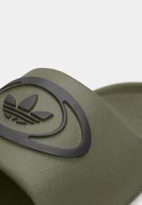 Zelená gumová šľapka s textúrovaným povrchom, na ktorej je výrazné čierne logo. Dizajn je jednoduchý a obsahuje zakrivené akcenty.