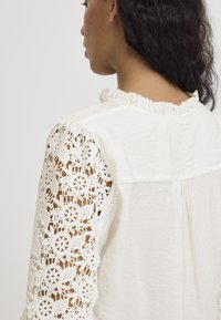 Blusa bianca con colletto arricciato, corpo dal texture, e maniche in pizzo intricate con motivi floreali e dettagli a intaglio.