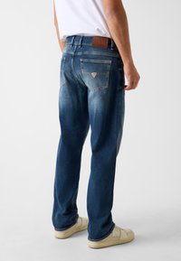 Jeans in denim blu scuro con finitura slavata, caratterizzati da una patch in pelle con il logo Guess sulla cintura e dettagli distintivi di cucitura sulle tasche posteriori.