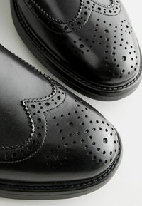 Chaussures en cuir noir au design brogue avec des détails perforés, des bords festonnés et un bout arrondi. Texture lisse avec des accents cousus.