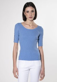 Top de punto azul con mangas cortas, que presenta un diseño de ribete texturizado y detalles de botones en los puños. Se lleva con pantalones blancos ajustados.