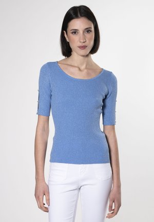 Top blu in maglia con maniche corte, caratterizzato da un design a coste testurizzate e inserti con bottoni sui polsini. Indossato con pantaloni bianchi aderenti.