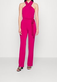 Fuchsia jumpsuit met een halternek ontwerp en een strikdetail bij de taille, gemaakt van een zachte stof, met wijd uitlopende broekspijpen en zwarte sandalen.