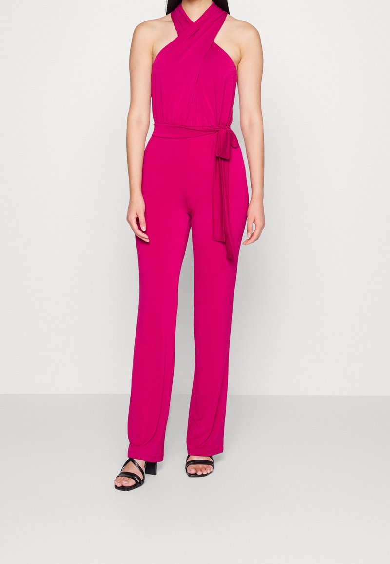 Fuchsia jumpsuit met een halternek ontwerp en een strikdetail bij de taille, gemaakt van een zachte stof, met wijd uitlopende broekspijpen en zwarte sandalen.