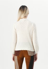 Frau mit langen blonden Haaren trägt einen weißen Strickpullover und eine gepatchte braune Cordhose, steht mit dem Rücken zu einem weißen Hintergrund.