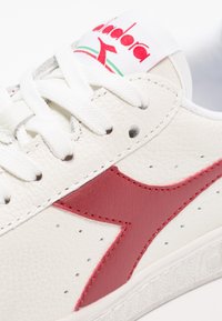Sneaker in pelle bianca con accenti rossi. Presenta una tomaia texture, punta rotonda, perforazioni e lacci bianchi piatti. Branding sulla linguetta.