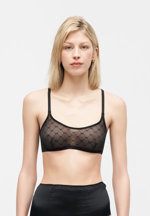 Jeune femme aux cheveux blonds portant un soutien-gorge noir transparent à motifs et un bas noir taille haute, sur un fond clair uni.