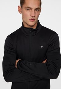 Svart zip-up jacka med hög krage, tillverkad av slätt tyg. Har raglanärmar och en subtil logga på bröstet och ärmen.