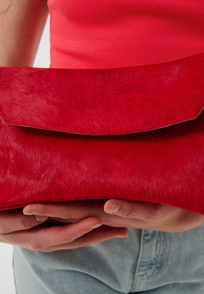 Red fur clutch bag with a smooth texture, fold-over flap, and no visible hardware. Held between hands, with denim fabric visible in the background.