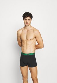 Schwarze Boxershorts mit grünem elastischen Bund, auf dem das "PUMA"-Logo abgebildet ist. Das Material wirkt glatt, die körpernahe Passform betont den Oberkörper.