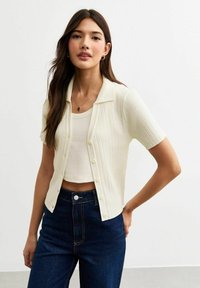 Korte mouwen, geribbelde cardigan in lichtbeige, met een kraag en knopen aan de voorkant, gecombineerd met een zacht wit tanktop en donkere jeans.