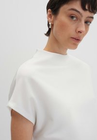 Haut blanc à manches courtes avec un col haut et une texture lisse, présentant des poignets roulés et un design minimaliste.