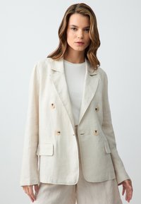 Leinen-Doppelreiher-Blazer in Hellbeige mit zwei großen Knöpfen, Reverskragen und vorderen Taschen, kombiniert mit einem weißen Hemd.