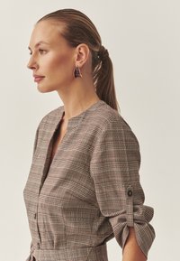 Barna kockás blúz v-nyakkal, gombos ujjakkal és megkötős derékkal. A modellen lila geometrikus fülbevalók vannak. Sima textúra.