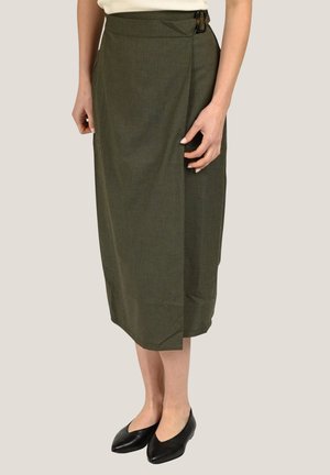 Femme portant une jupe midi taille haute vert olive avec une boucle sur le côté, associée à des ballerines pointues noires et un haut blanc.