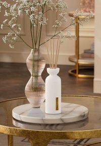 Hvit rillede keramikkdiffuser med bambusstaver ved siden av en rosa presset glassvase på et rundt marmorbrett på et gullkaffebord.