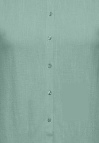 Gaiši tējas krāsas pogājama krekls ar garām piedurknēm, rotāts ar maziem apaļiem pogām, kas saskan ar audumu un izvietotas priekšējā atloka daļā.