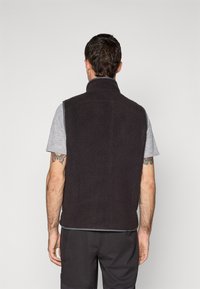 Man met kort bruin haar, die een zwart fleece vest over een grijs shirt met korte mouwen draagt, staande met zijn rug naar de camera tegen een effen achtergrond.