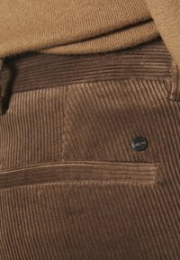 Un pantalon en velours côtelé marron présente une surface texturée, avec un design élégant. Un petit bouton en métal noir orne la poche arrière.
