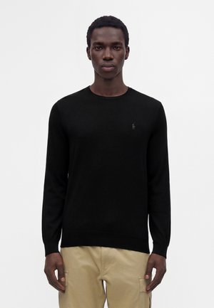 Homme portant un pull noir à manches longues et un pantalon beige, debout devant un fond blanc uni.