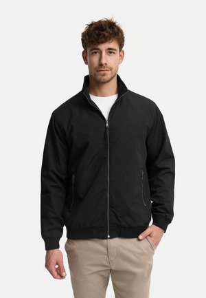 Młody mężczyzna ubrany w czarną kurtkę na zamek, białą koszulę i beżowe spodnie, stoi z jedną ręką w kieszeni na jednolitym tle.