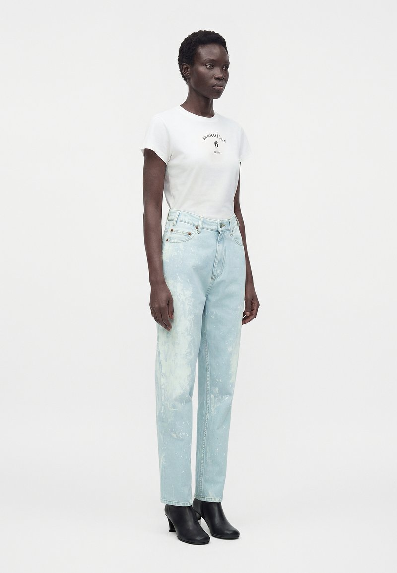 Vrouw met kort krullend haar, die een wit Margiela-logo T-shirt draagt, lichtblauwe gewassen rechte jeans en zwarte enkellaarzen met hak, staand zijwaarts.