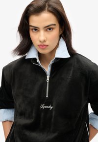 Felpa nera in velluto a coste con zip parziale e ricamo bianco "Superdry", indossata sopra una camicia azzurra a colletto. Tessuto morbido, vestibilità comoda.