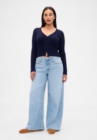 Temno modra rebrasta jopica z gumbi, kombinirana s svetlo modrimi širokimi kavbojkami. Model nosi rjave balerinke na enostavni belem ozadju.