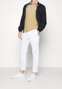 Černá bunda s kostkovanou podšívkou, béžové polo tričko, bílé zúžené kalhoty a bílé tenisky. Jednoduchý design s hladkými texturami.