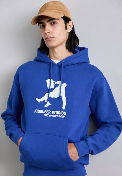 Felpa blu reale con cordini, tasca frontale e stampa grafica bianca che presenta una figura e del testo. Tessuto morbido, design casual.