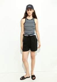 Striped tank top in navy and white, black drawstring shorts, and black slide sandals. Cap features logo detail. Smooth fabric textures.