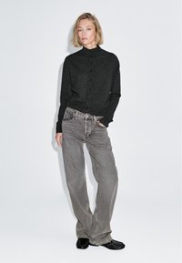 Cardigan nero con bottoni e collo alto, abbinato a jeans grigi larghi e scarpe nere, su uno sfondo bianco semplice.
