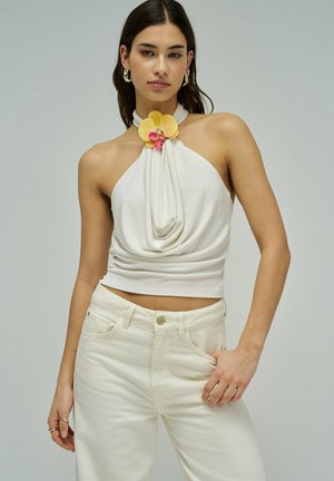 Joven mujer con top halter color crema con detalle de flor y pantalones crema de cintura alta, posando con una mano en el bolsillo contra un fondo gris.