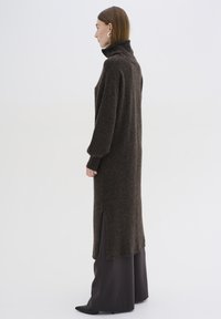 Hněd dress s pleteným vzorem, vysokým límcem, dlouhými rukávy a bočními rozparky. Materiál vypadá měkký a texturovaný, s uvolněným střihem.