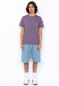T-shirt di cotone viola con collo rotondo e taschino a sinistra, abbinata a pantaloni shorts in denim. Il modello indossa calze bianche e sneakers.