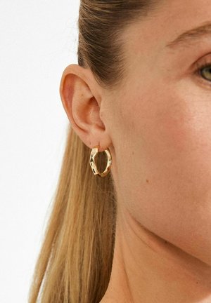 Kuldne kõrvarõngas lainelise disainiga, millel on sile pind ja poleeritud viimistlus. Kõrvarõngas on kantud osaliselt nähtavale kõrvale.