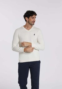 Pull en V crème à manches longues, arborant un petit emblème marine sur la poitrine, assorti à un pantalon bleu foncé. Texture lisse.