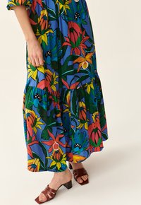 Blumenkleid im Maxiformat mit blauem Hintergrund, das lebendige rote, gelbe und grüne Muster zeigt. Mit gestuftem Saum und kurzen Ärmeln, kombiniert mit braunen Riemchen-Sandalen.