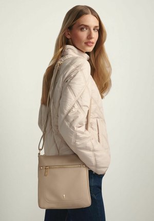 Kobieta z długimi blond włosami, ubrana w pikowaną jasnobeżową kurtkę i niebieskie dżinsy, nosząca taupe skórzaną torbę na ramię z złotym suwakiem.