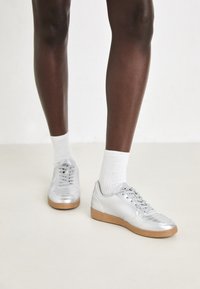 Silberne Sportschuhe mit einer glatten synthetischen Oberseite, Gummisohle und Schnürdesign, kombiniert mit weißen Kniestrümpfen.