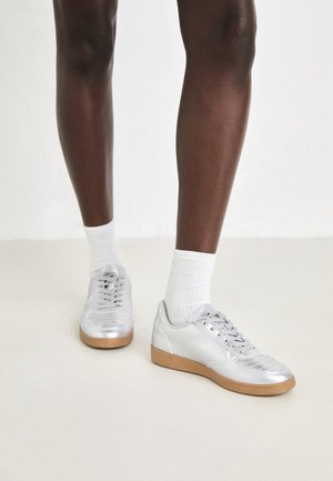 Sølv idrettssko med en glatt syntetisk overdel, gummisåle og snøring, kombinert med hvite ankelstrømper.