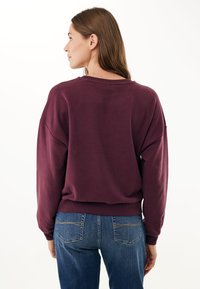Felpa oversized bordeaux con spalle scese, polsini a coste e scollatura rotonda, abbinata a jeans blu con tasche posteriori.