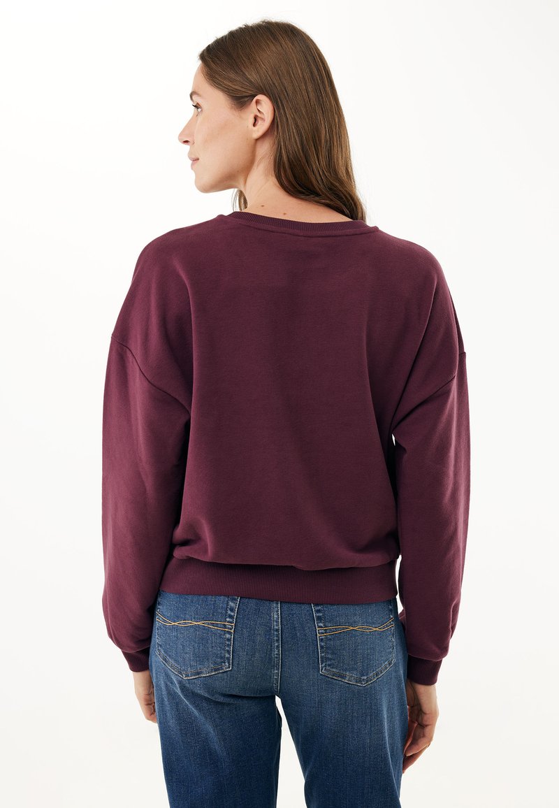 Bordeaux oversized sweatshirt met verlaagde schouders, geribbelde boorden en een ronde halslijn, gecombineerd met blauwe jeans met achterzakken.