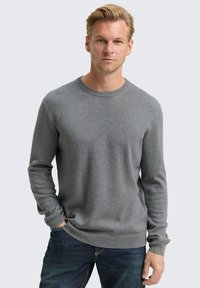 Blonde man in een effen grijze sweater met lange mouwen en donkere spijkerbroeken, met één hand in zijn zak, recht vooruit kijkend tegen een lichte achtergrond.
